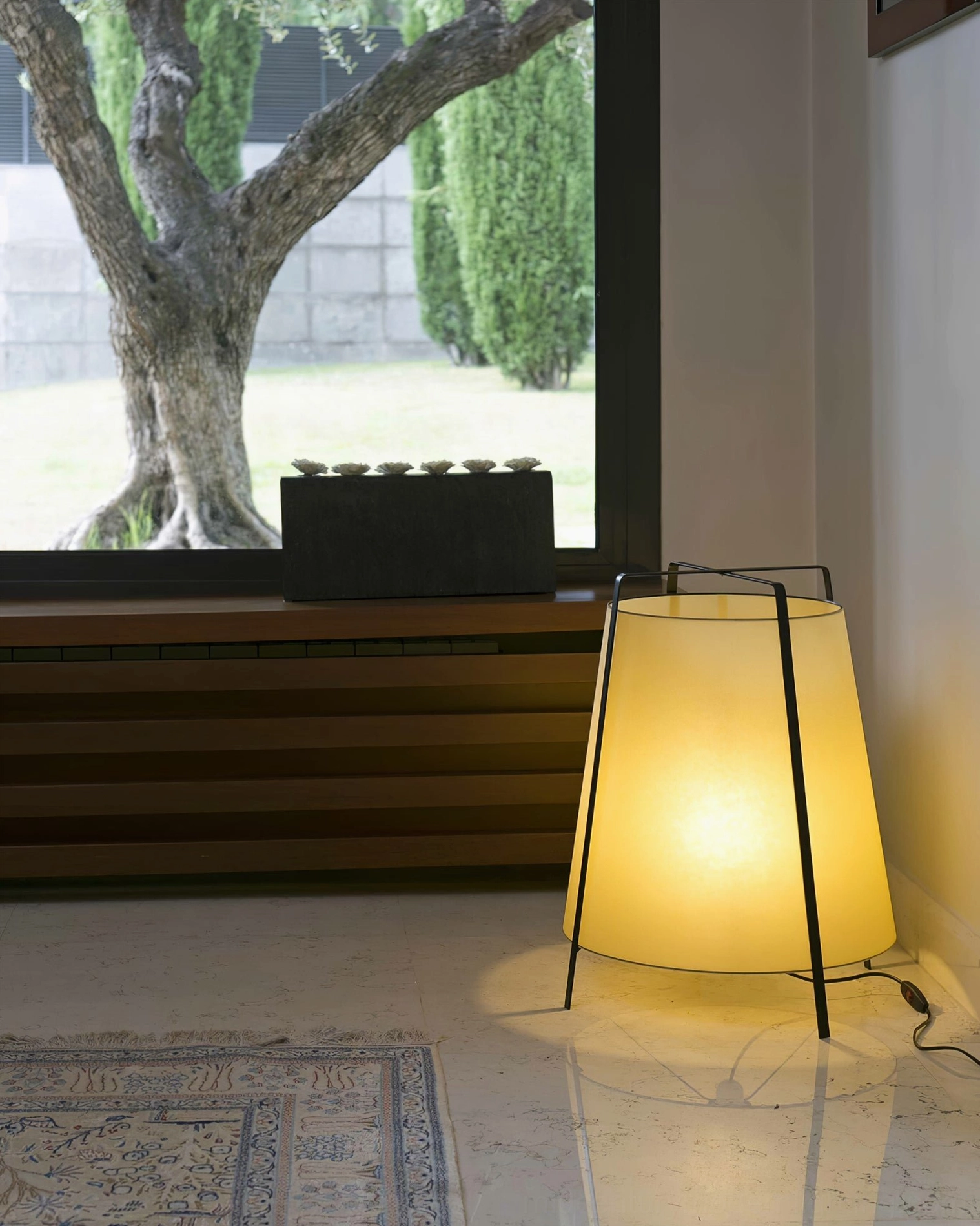 Akane table lamp by window with marble floor and outdoor tree view, side three-quarter angle showing warm light