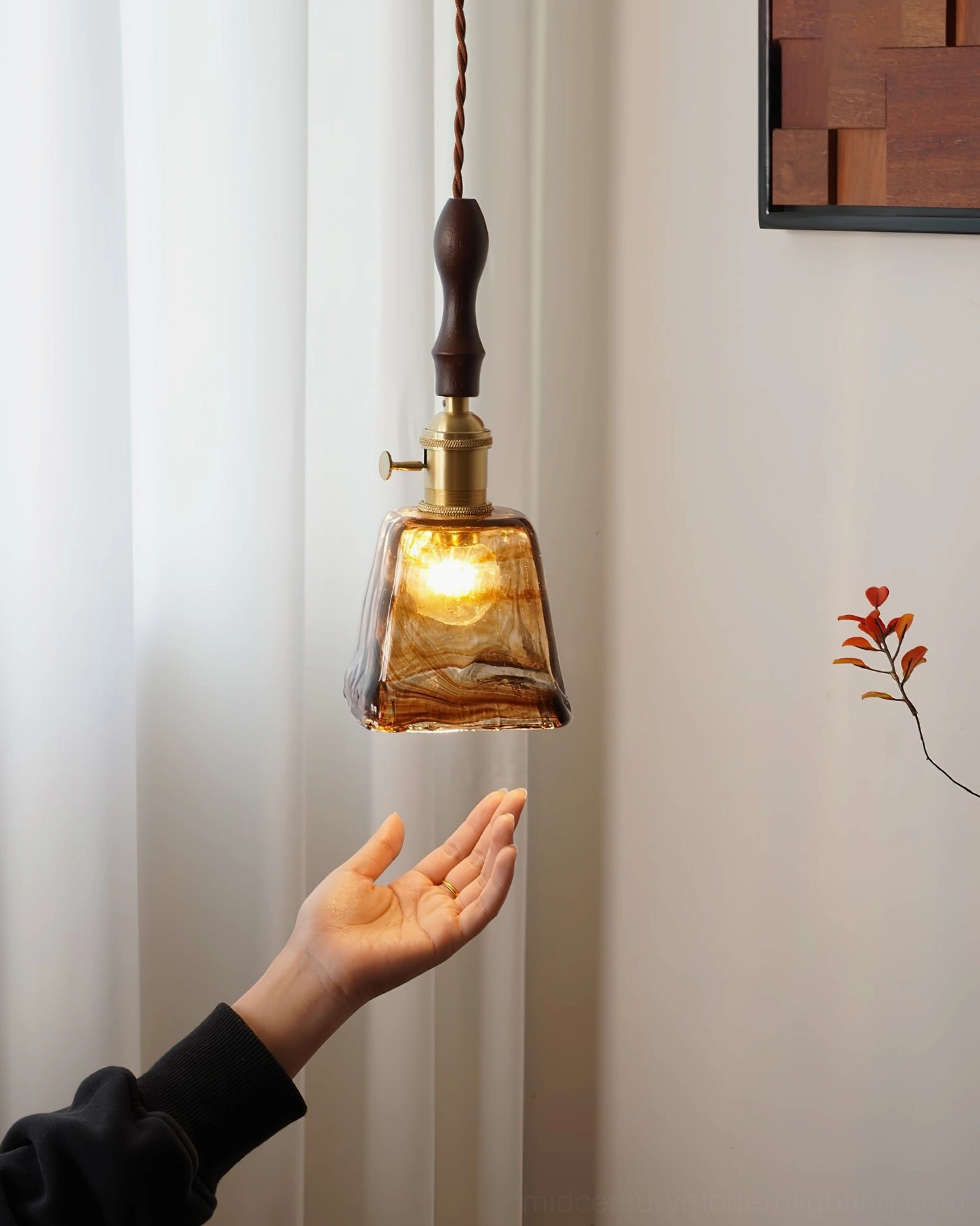 Hand holding sculptural amber glass pendant showing organic curves and walnut wood handle in residential corner setting