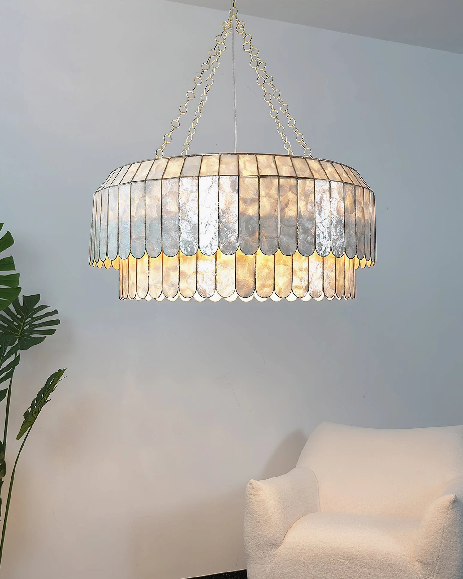 Two-tier capiz pendant lighting in cozy minimalist living room with white armchair and potted plants, sophisticated coastal luxury ambiance