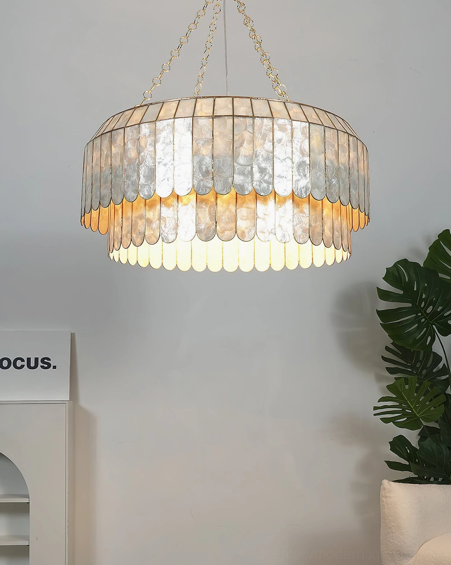 Two-tier capiz chandelier with warm glowing light in minimalist space with greenery and signage, low-angle frontal view, boho coastal luxury