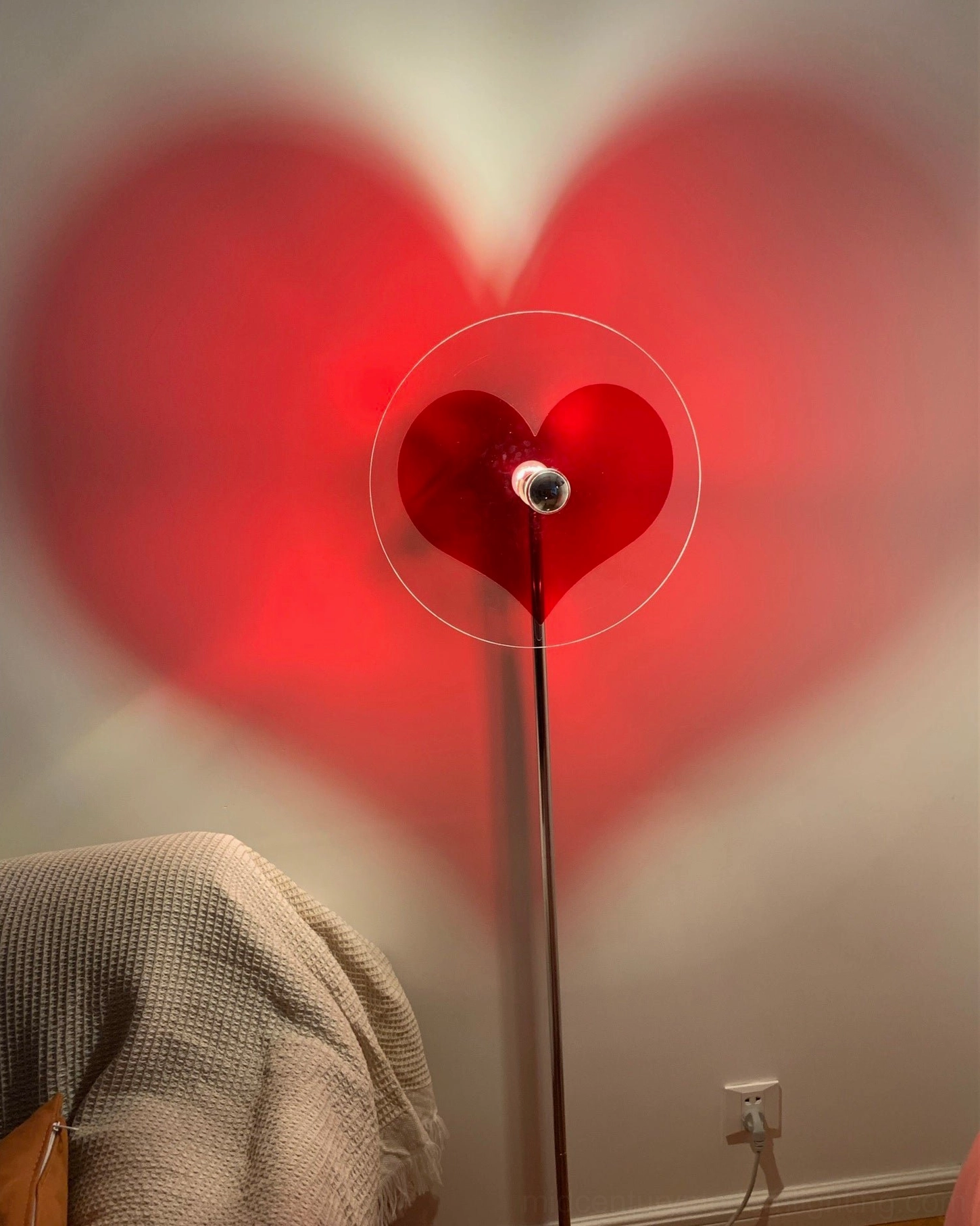 Heart floor lamp in bedroom corner with large soft red heart shadow near wall outlet, clear acrylic diffuser and chrome pole