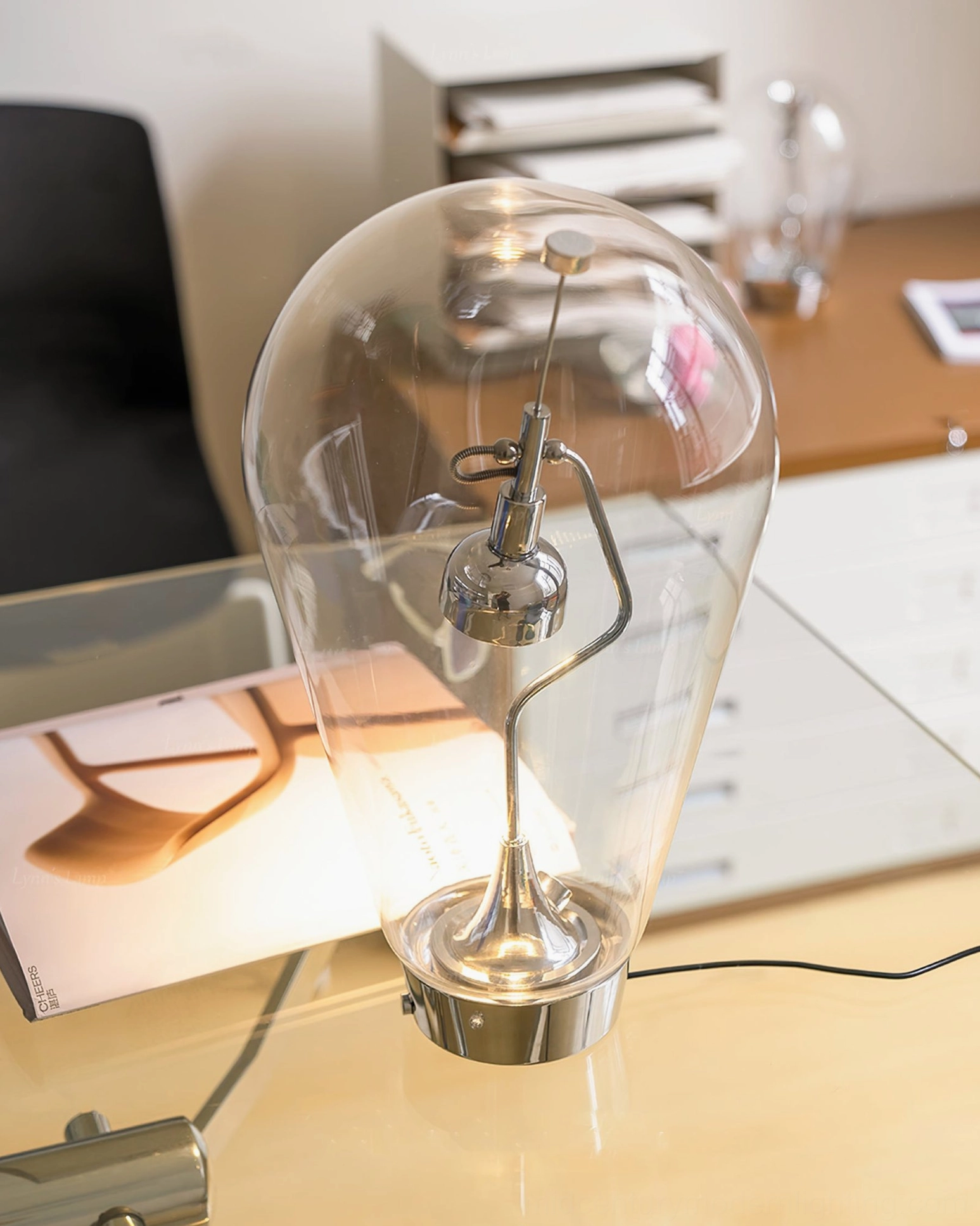 Blow illuminated table lamp three-quarter front view glowing on modern glass desk with magazine - warm white LED light