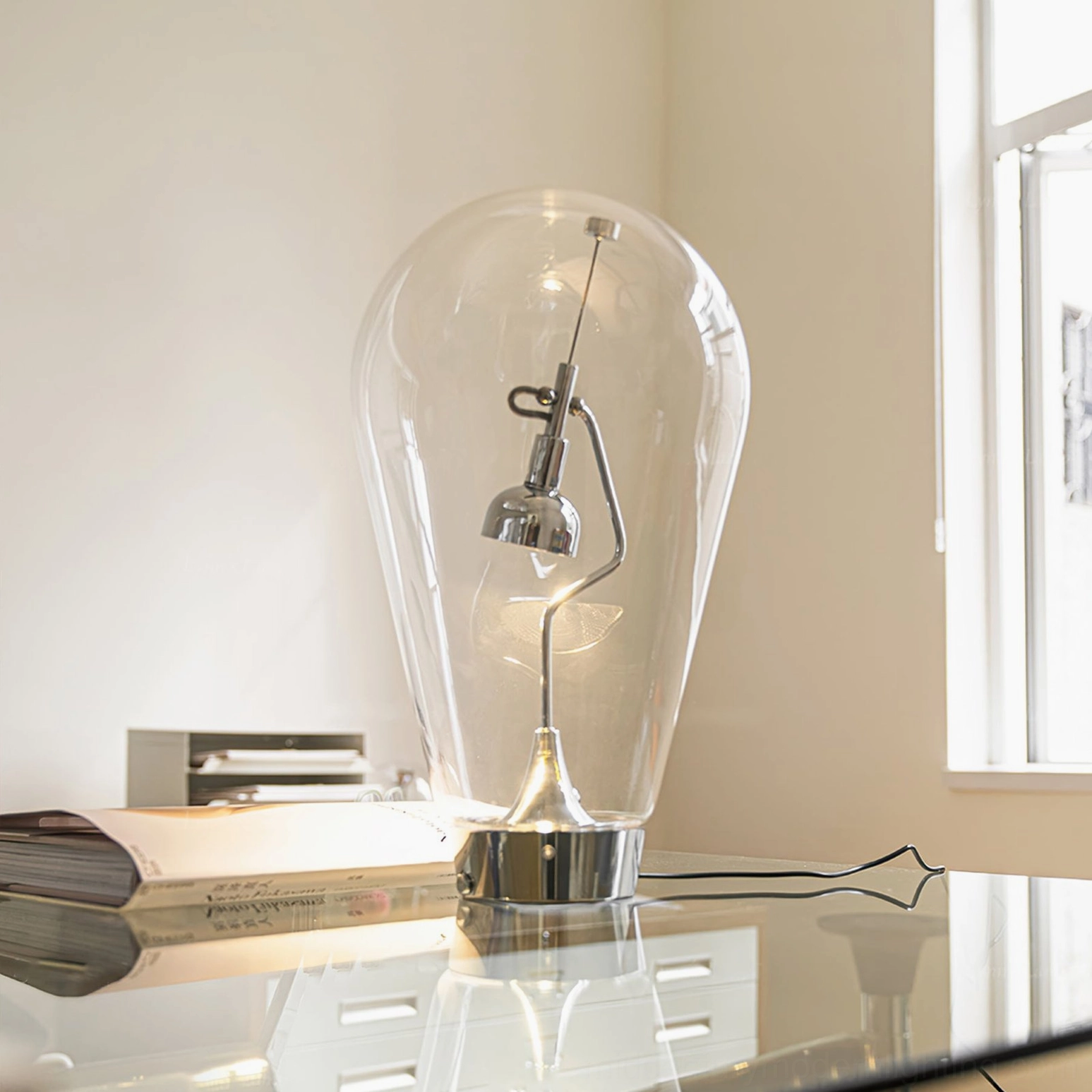 Blow modern table lamp three-quarter side view on glass desk with stacked books showing reflective glass shade and chrome base
