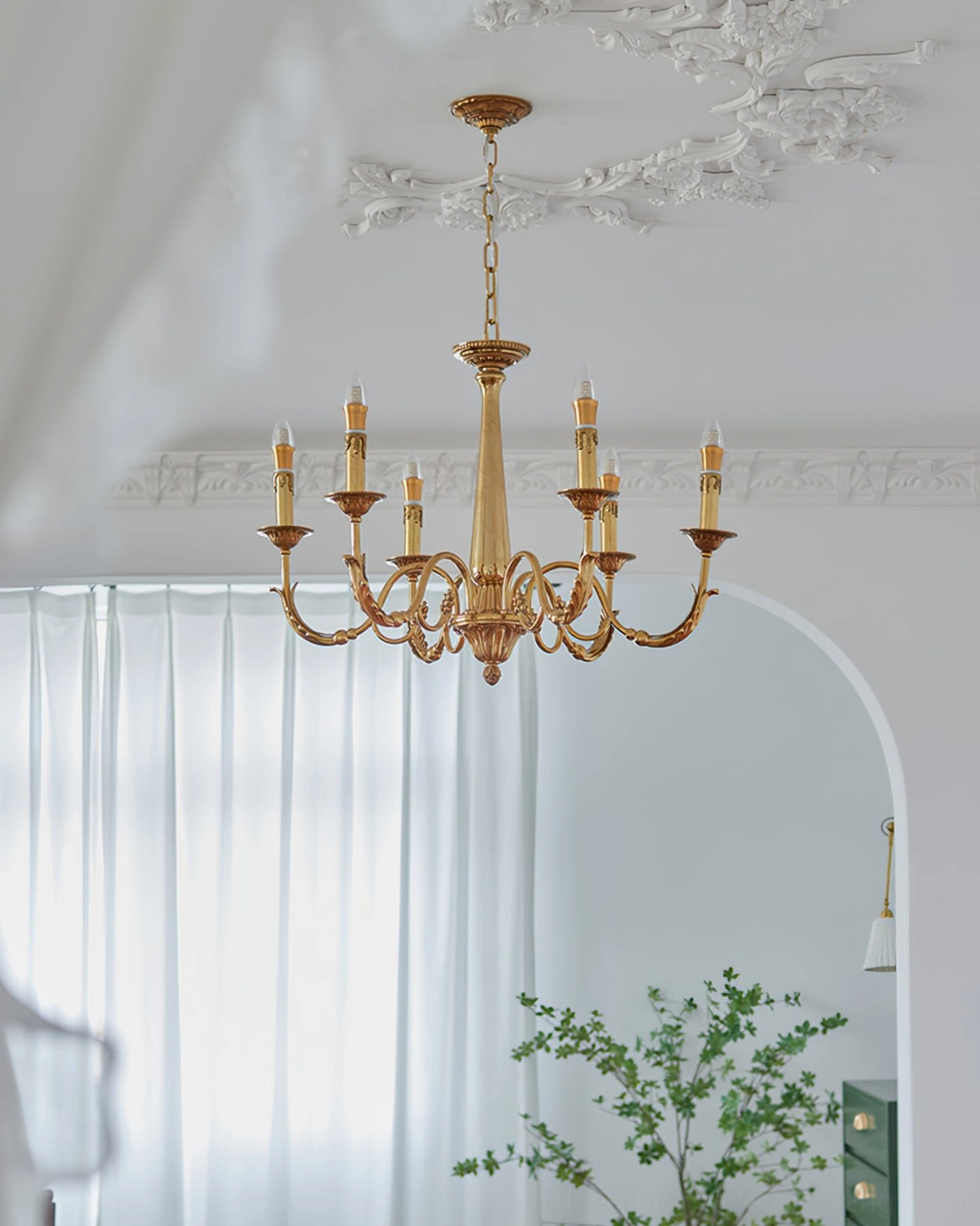 Brass candle chandelier installed in bright bedroom with sheer white curtains and arched mirror