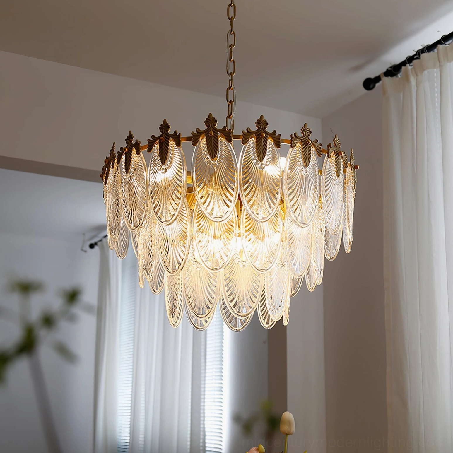 Illuminated three-ring brass chandelier in living room with sheer curtains, displaying warm light diffused through opaque glass leaves