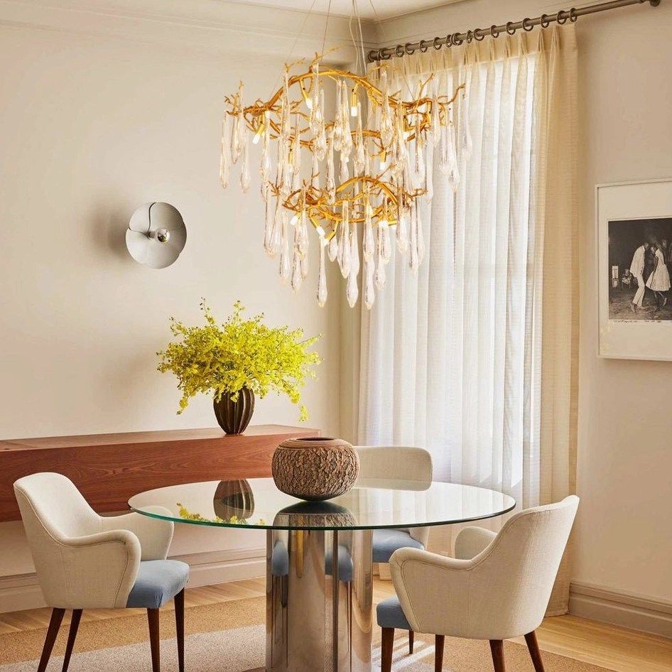 Modern dining room interior featuring brass waterfall chandelier with hanging crystal glass pendants above glass dining table with warm ambient lighting