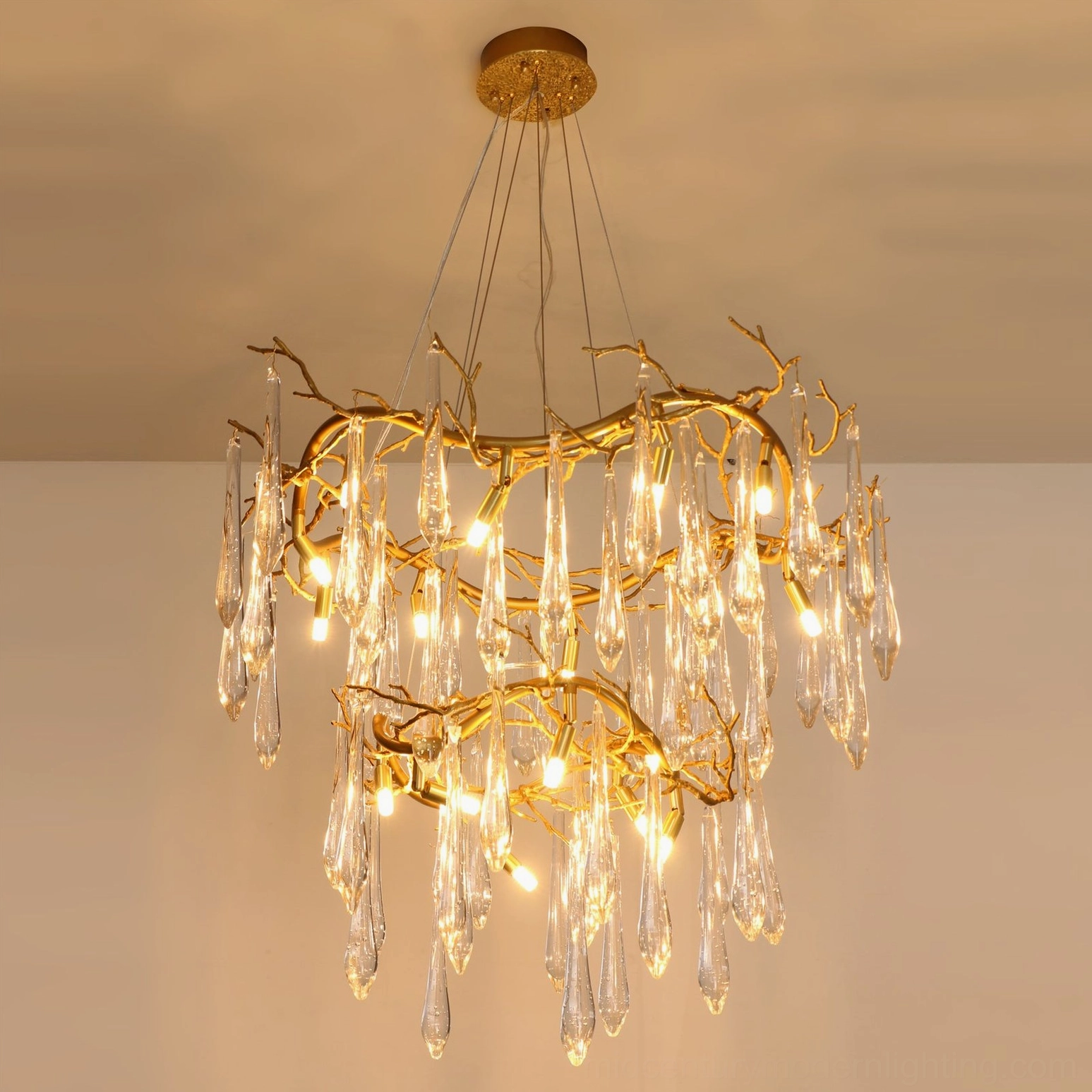 Fully illuminated brass waterfall chandelier with warm gold and clear glass aesthetic, slanted overhead view against warm indoor wall background