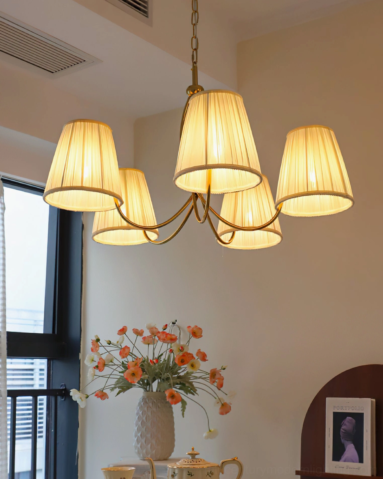 Lit Calistoga 5-light chandelier side view, glowing warm light in living area with black window frame and orange flowers