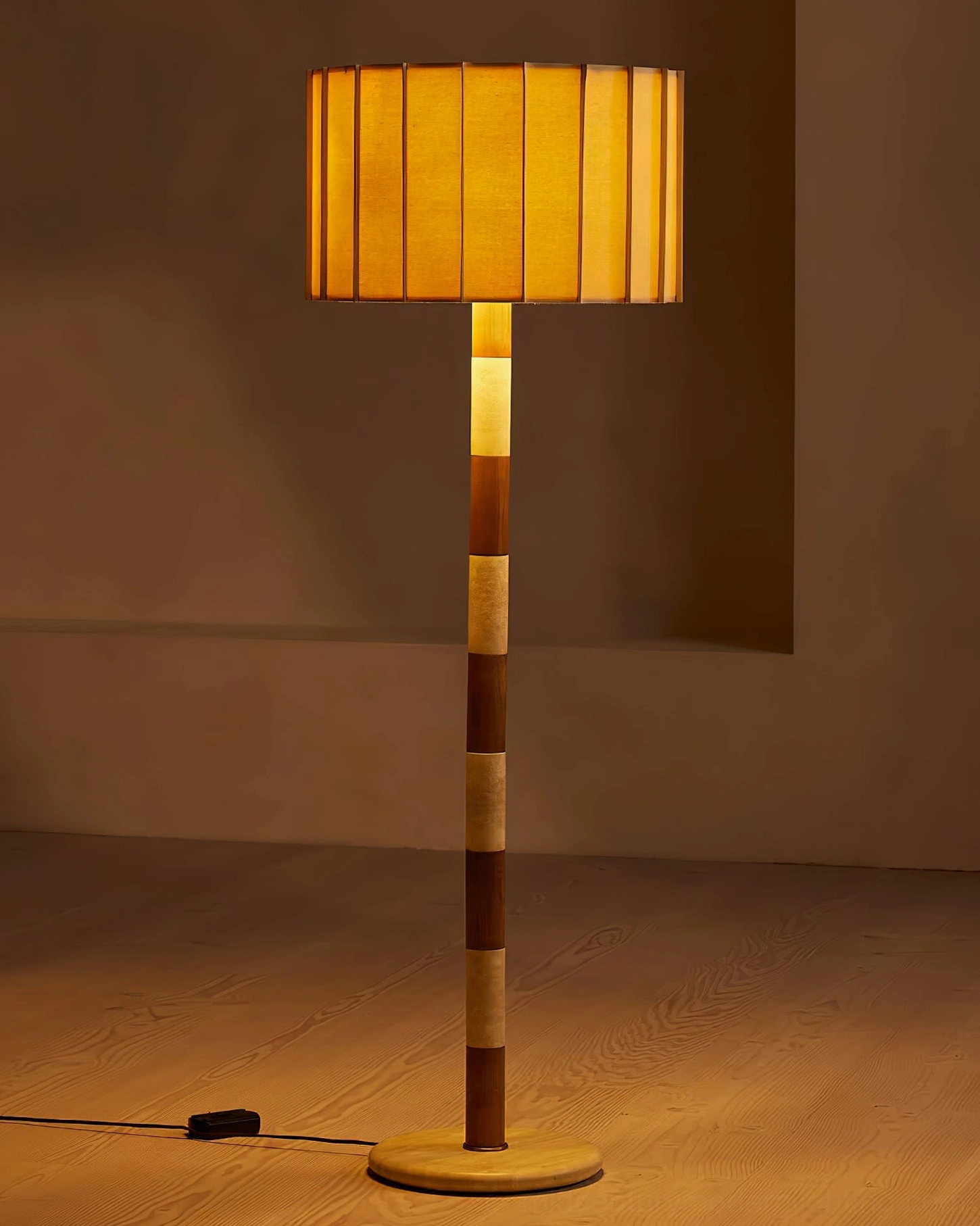 Illuminated Calypso floor lamp with glowing pleated shade and brass metal base on warm wooden floor, ambient lighting
