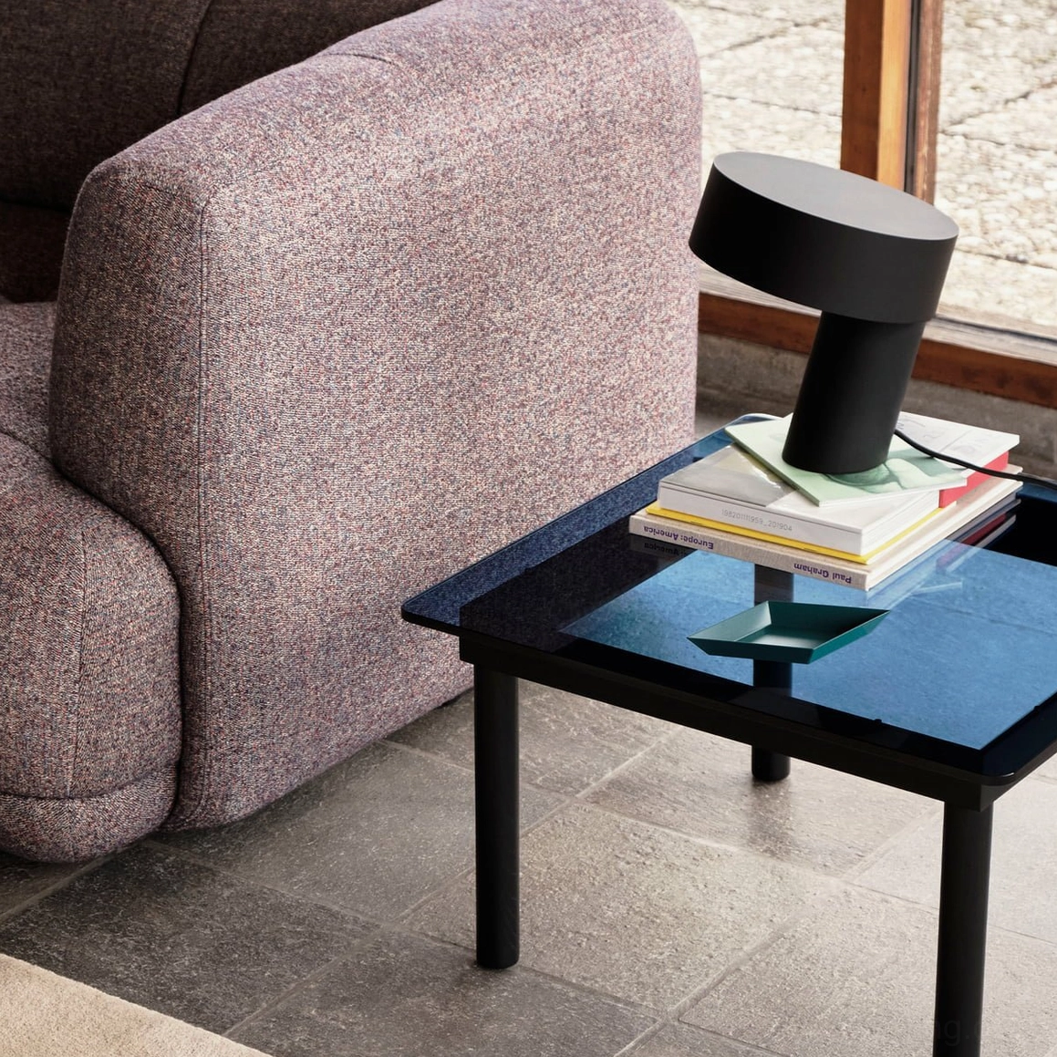 Matte black Celia angular lamp on glass side table next to gray fabric sofa with stacked books, modern interior