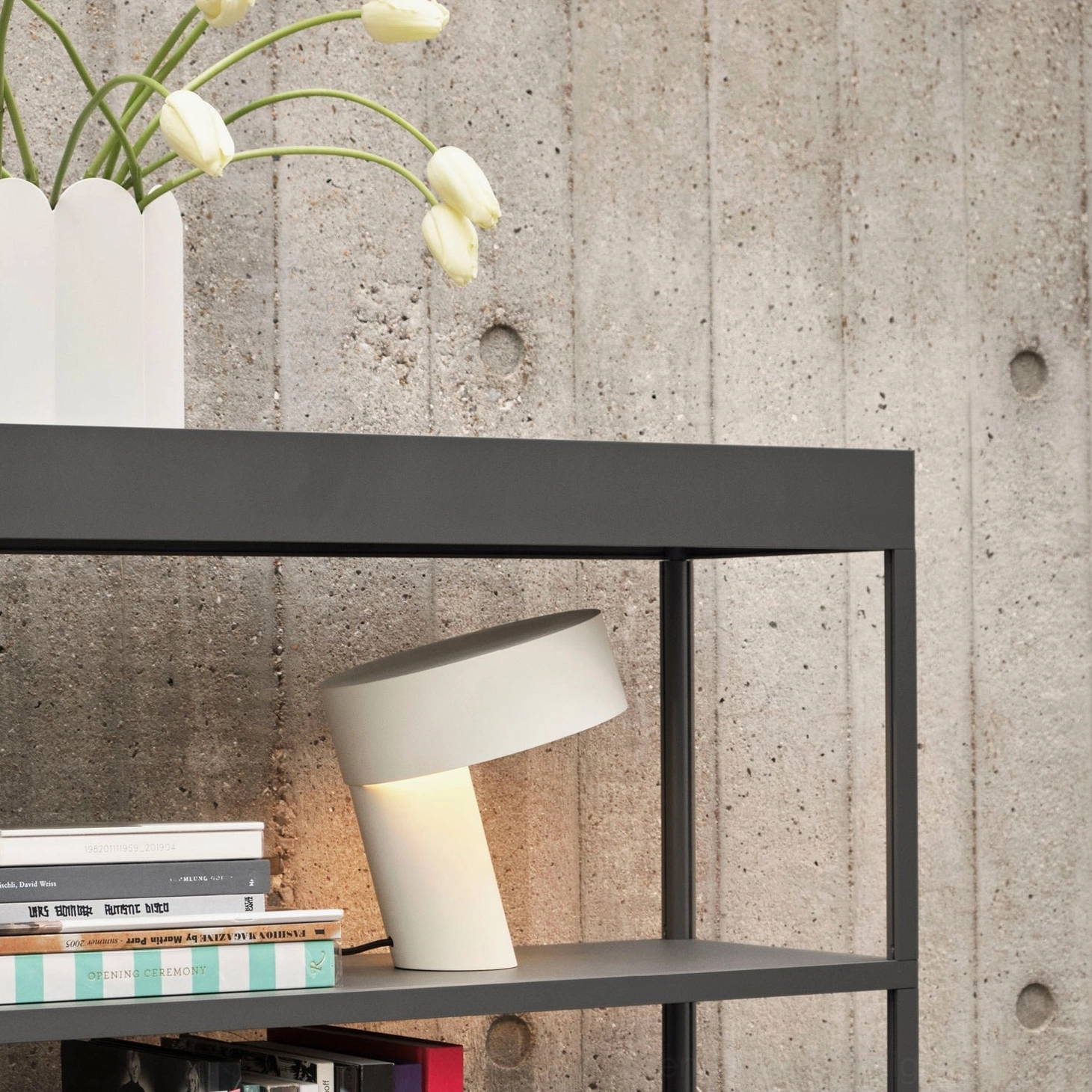Off-white Celia table lamp on metal shelf against concrete wall with books and white tulips, minimalist design