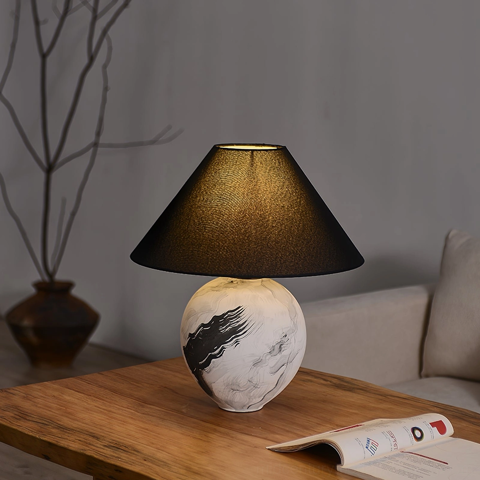Lit ceramic table lamp with illuminated linen shade on living room wooden table with sofa and plant decor