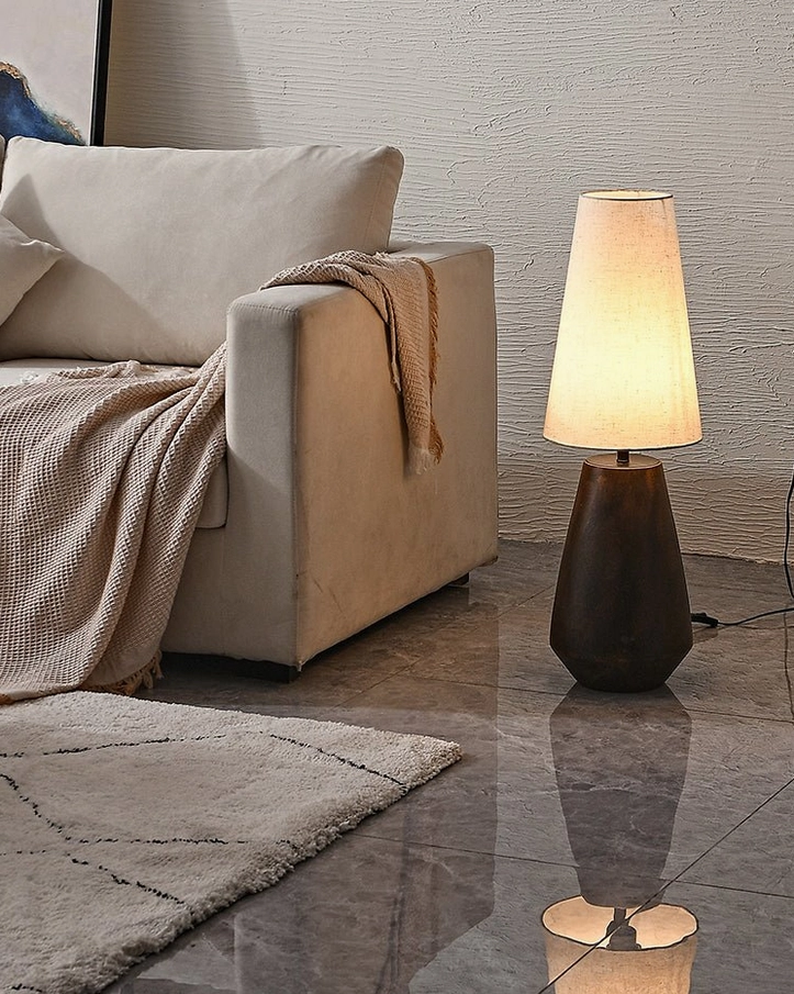 Low-angle view of ceramic lamp with reflection on tile floor, light-colored sofa with textile throw, showcasing ambient home lighting design