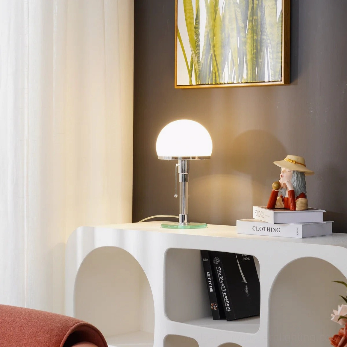 Hemispherical chrome lamp lit on living room console table with stacked books, decorative objects and sheer window drapes, warm ambient lighting