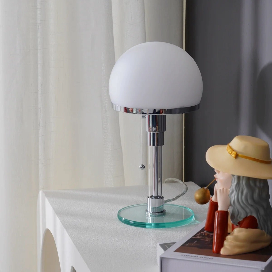 Chrome hemispherical table lamp glowing warmly on bedroom side table with decorative figurine, sheer curtain and cozy mid-century modern aesthetic