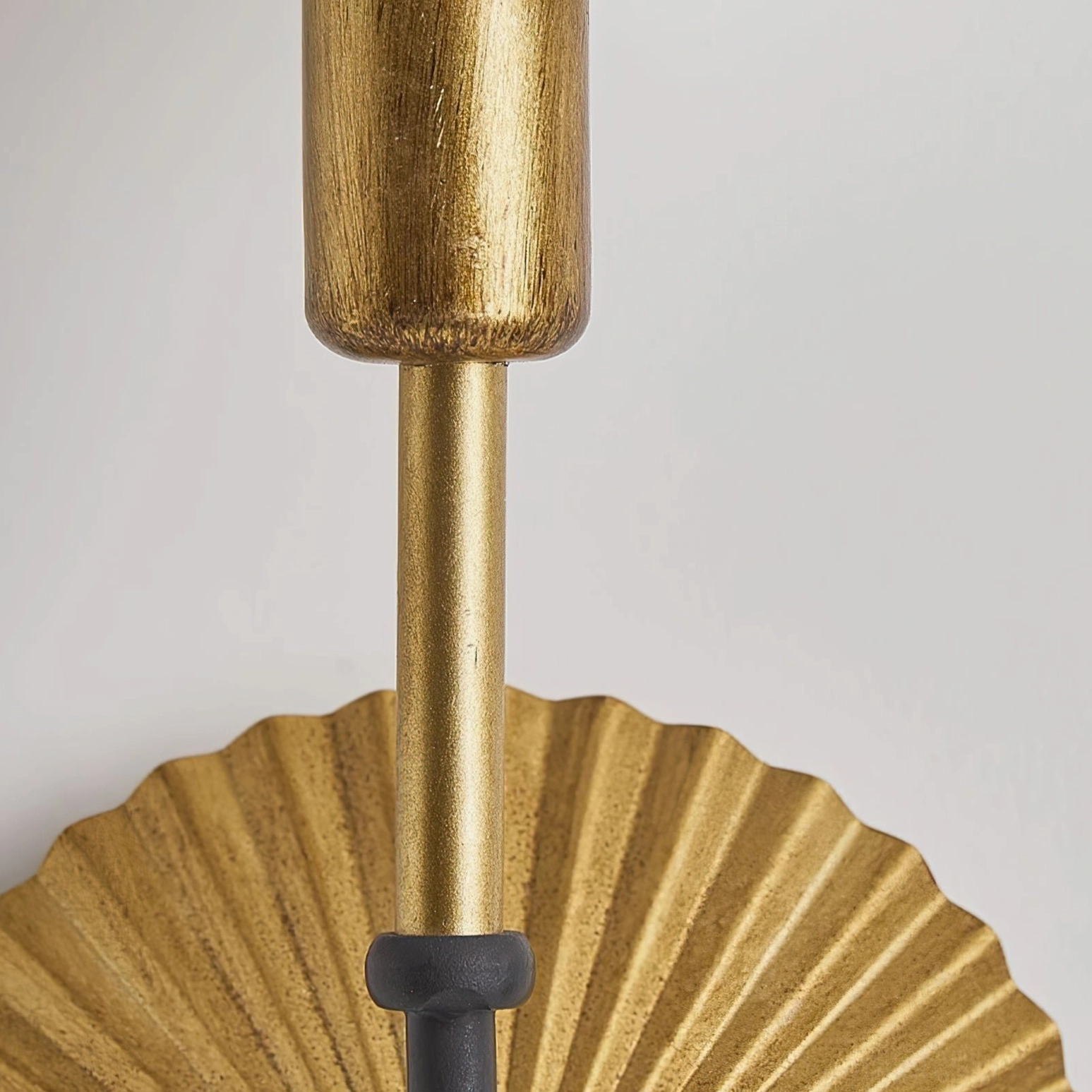 Low-angle close-up detail of Clove wall lamp showing brushed gold metal rod, pleated fan base structure, and matte black joint hardware