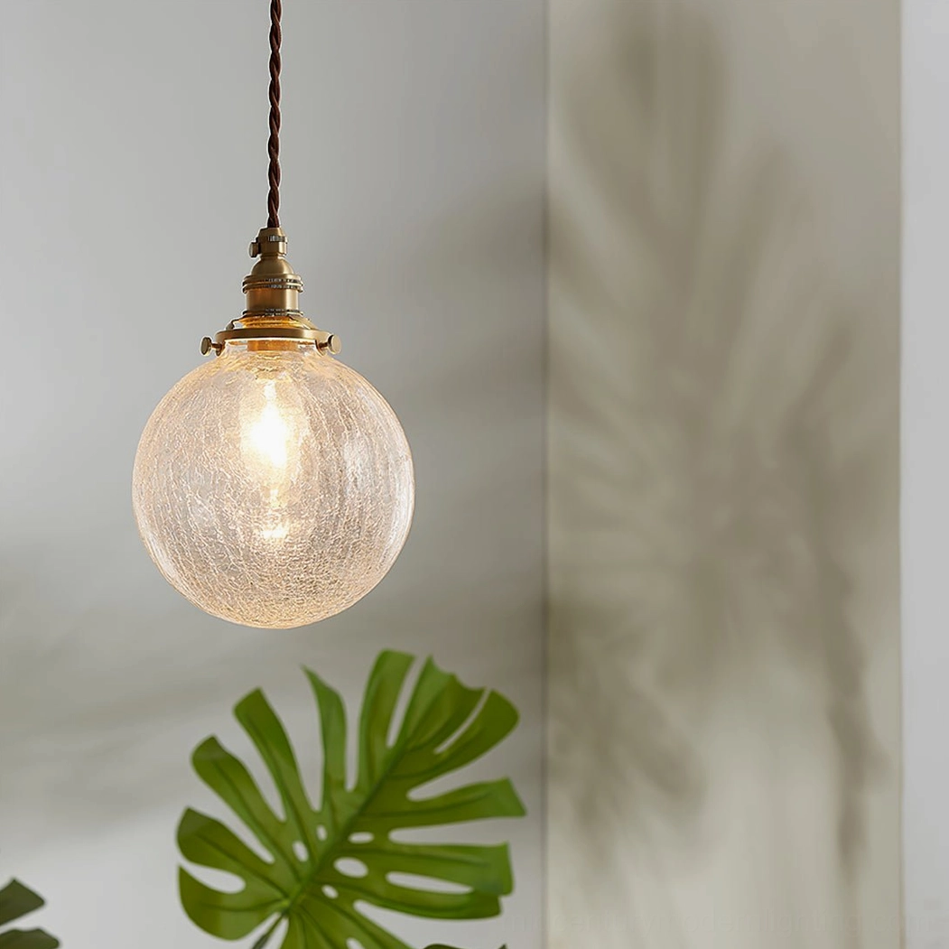 Close-up view of crackle glass pendant with twisted brass cord, textured glass surface detail, indoor corner with green foliage