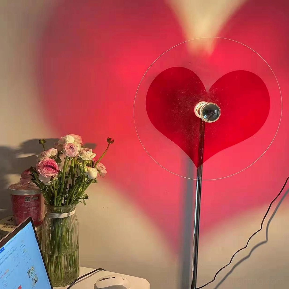 Heart-shaped floor lamp with red shadow projection on work desk, flower bouquet and laptop visible, power cord shown
