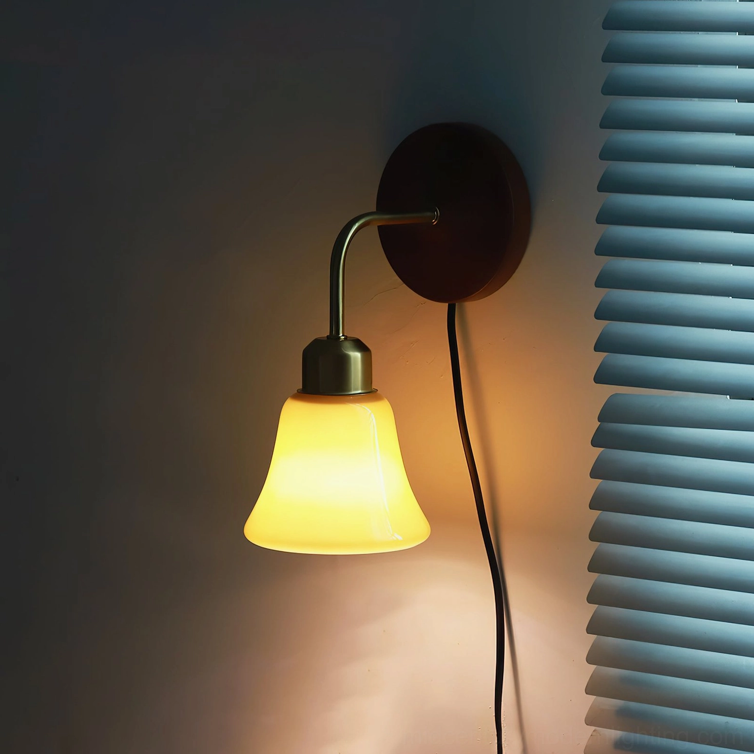 Daphne Pastel Wall Sconce illuminated side view casting warm yellow ambient glow against dark blue louver blinds, demonstrating soft lighting quality