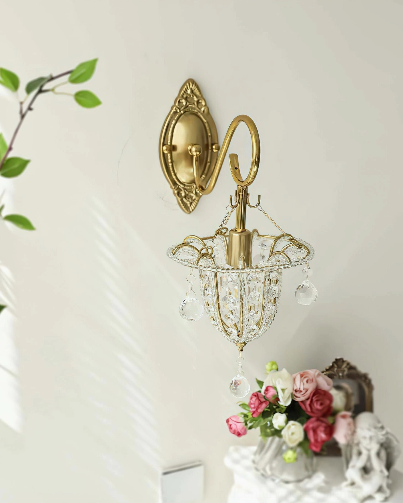 Front side view of Eilers ornate brass wall sconce mounted on white wall with green foliage and white flowers, displaying French country luxury aesthetic