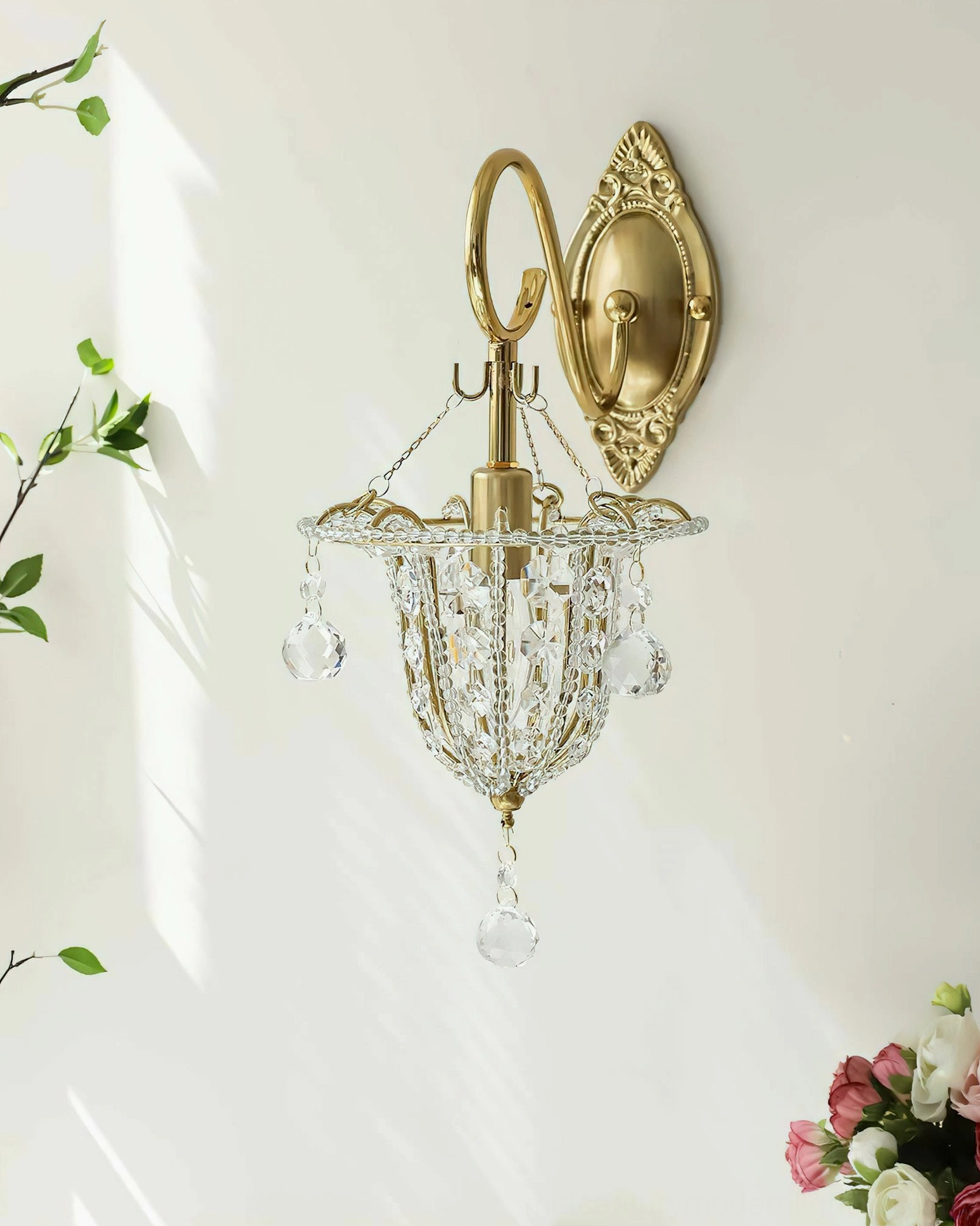 Eilers timeless crystal wall sconce in front flat view on sunlit white wall with trailing green vines, showing full profile of gold hardware and hanging crystal pendants