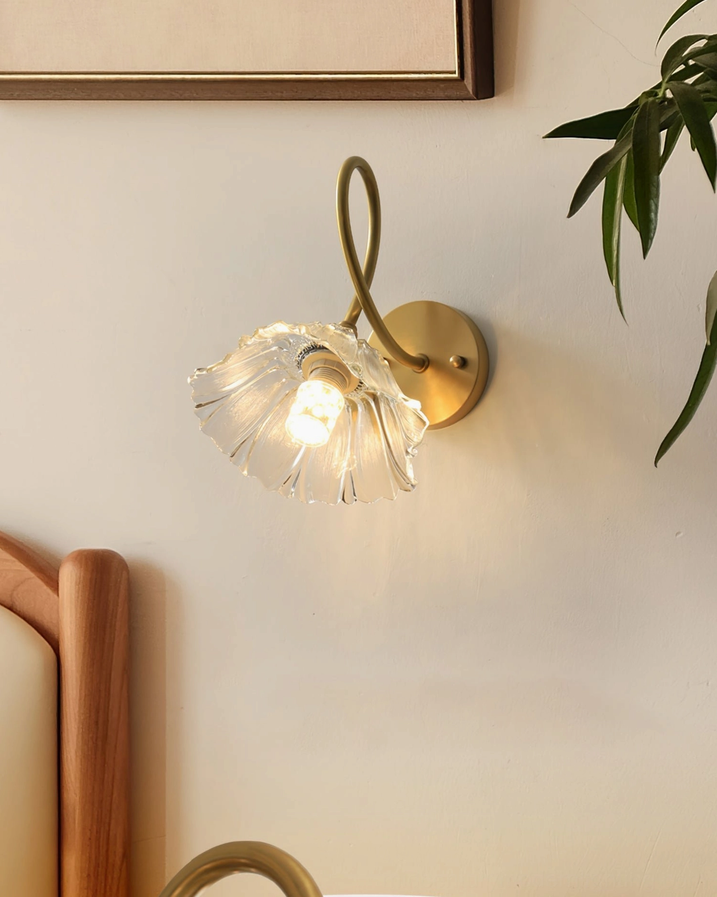 Ercole brass sconce in bedroom setting beside wooden bed frame with green potted plant