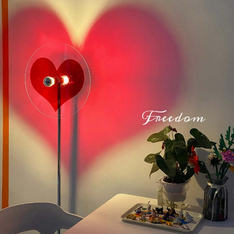 Red heart floor lamp creating large shadow projection with Freedom text on home office wall, potted plant decor visible