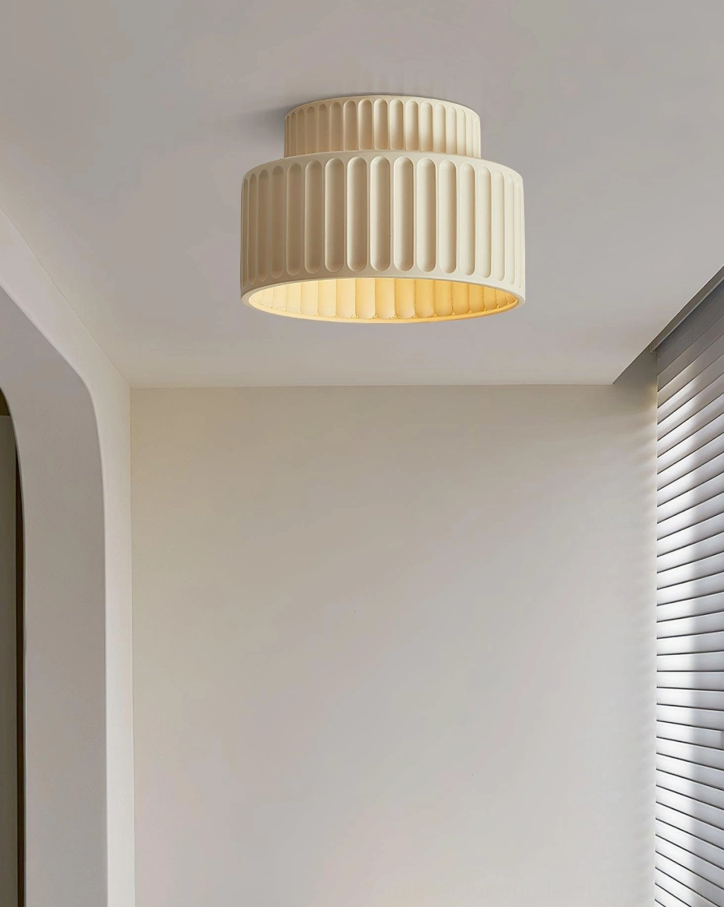 Beige Kami fluted ceiling lamp overhead front angle in minimalist room with louvered window shade, soft warm lighting
