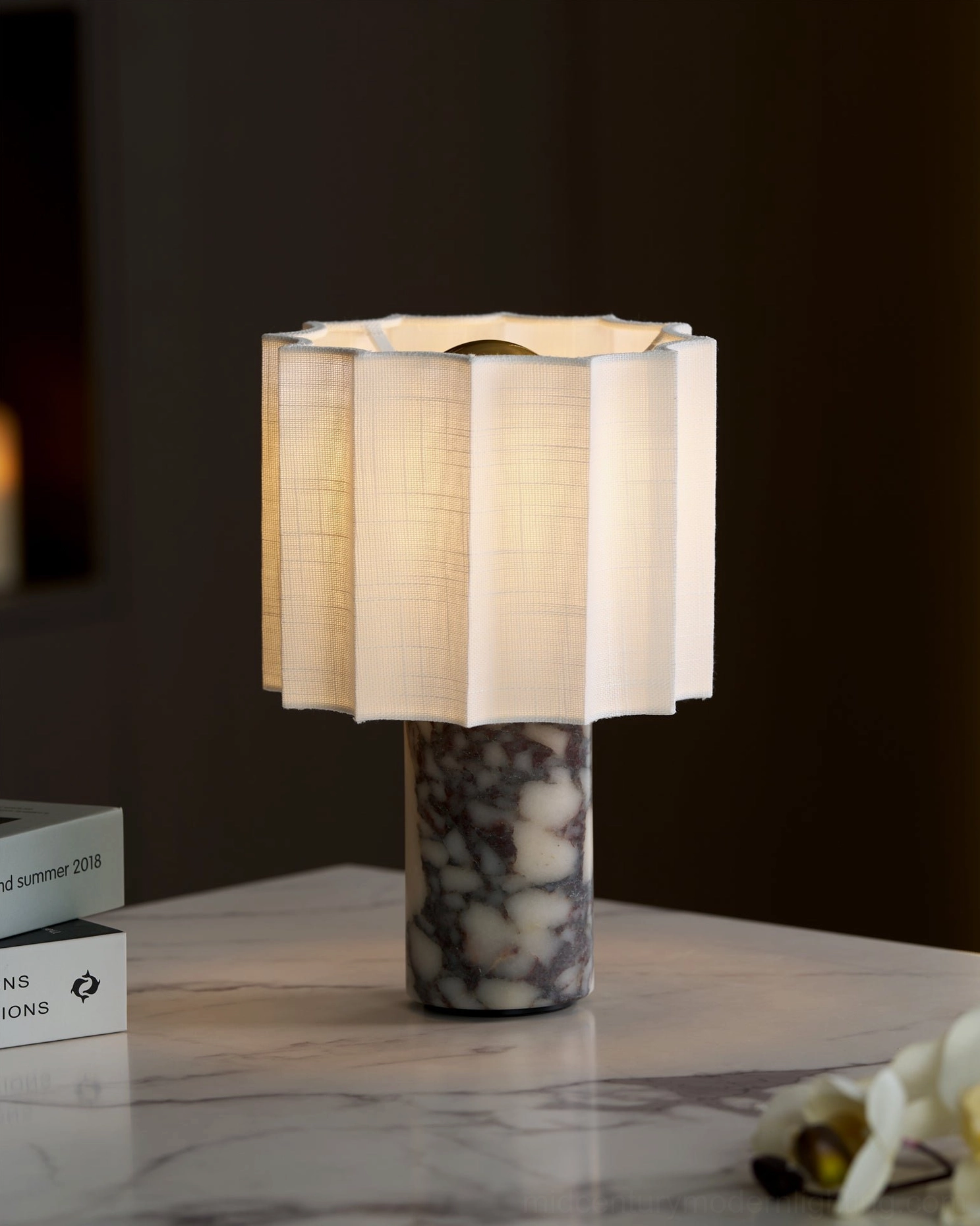 Glowing Kane cordless lamp on marble table from low three-quarter angle, warm-lit beige shade with dark marble base and decorative accents