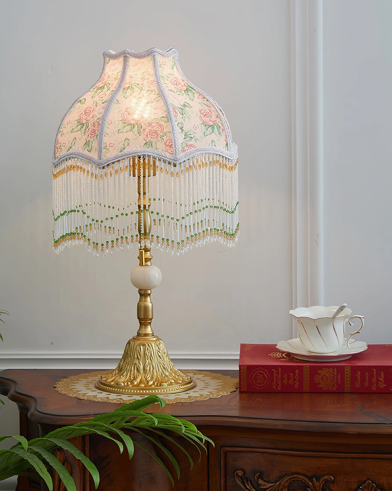 Masa lit beaded table lamp illuminated showing pink rose print fabric shade with green tassel and gold metal base glowing