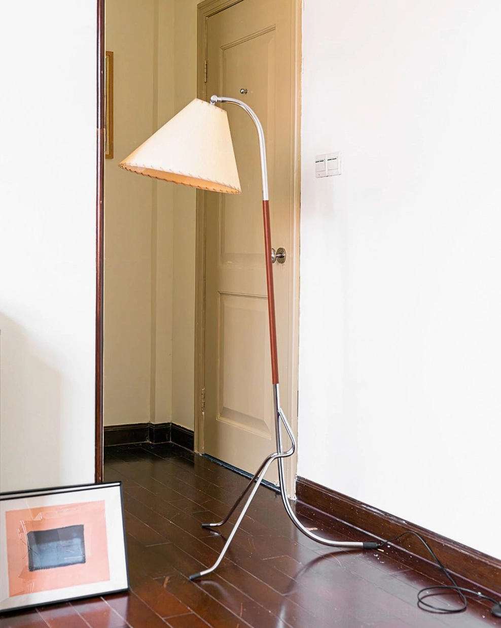 Mid-century tripod floor lamp standing in wooden hallway near door and framed artwork, showcasing interior design versatility