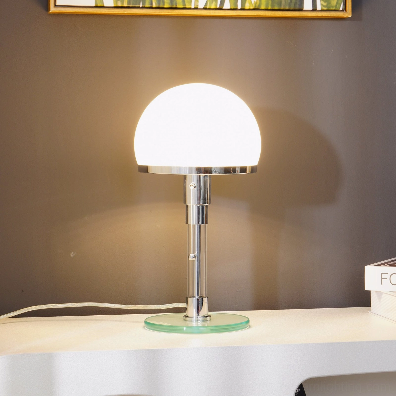 Modern chrome table lamp glowing on minimalist console table against dark gray wall with framed artwork, contemporary interior styling