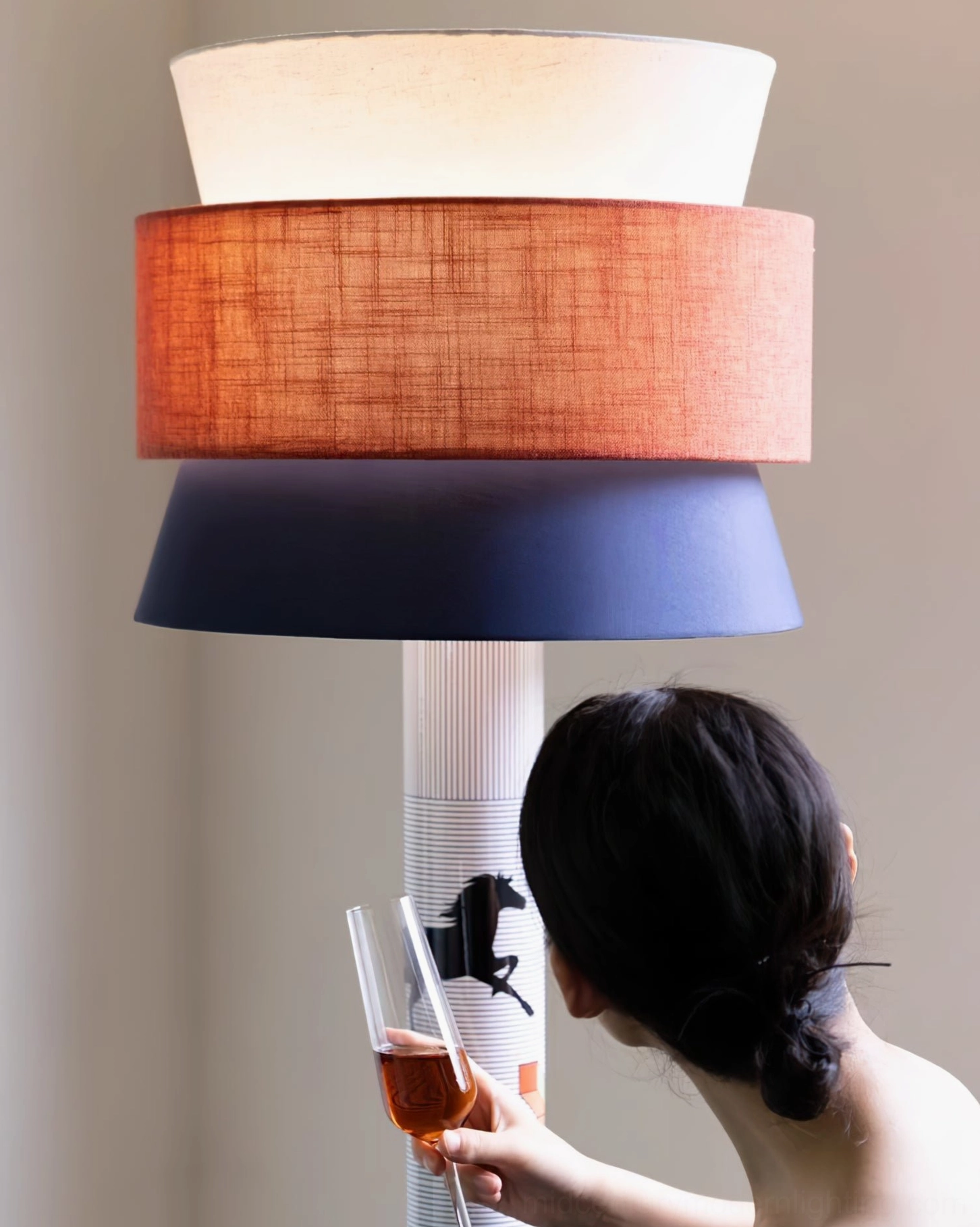 Artistic floor lamp close-up studio shot showing layered tri-color shade and equine printed metal pole with hand holding wine glass