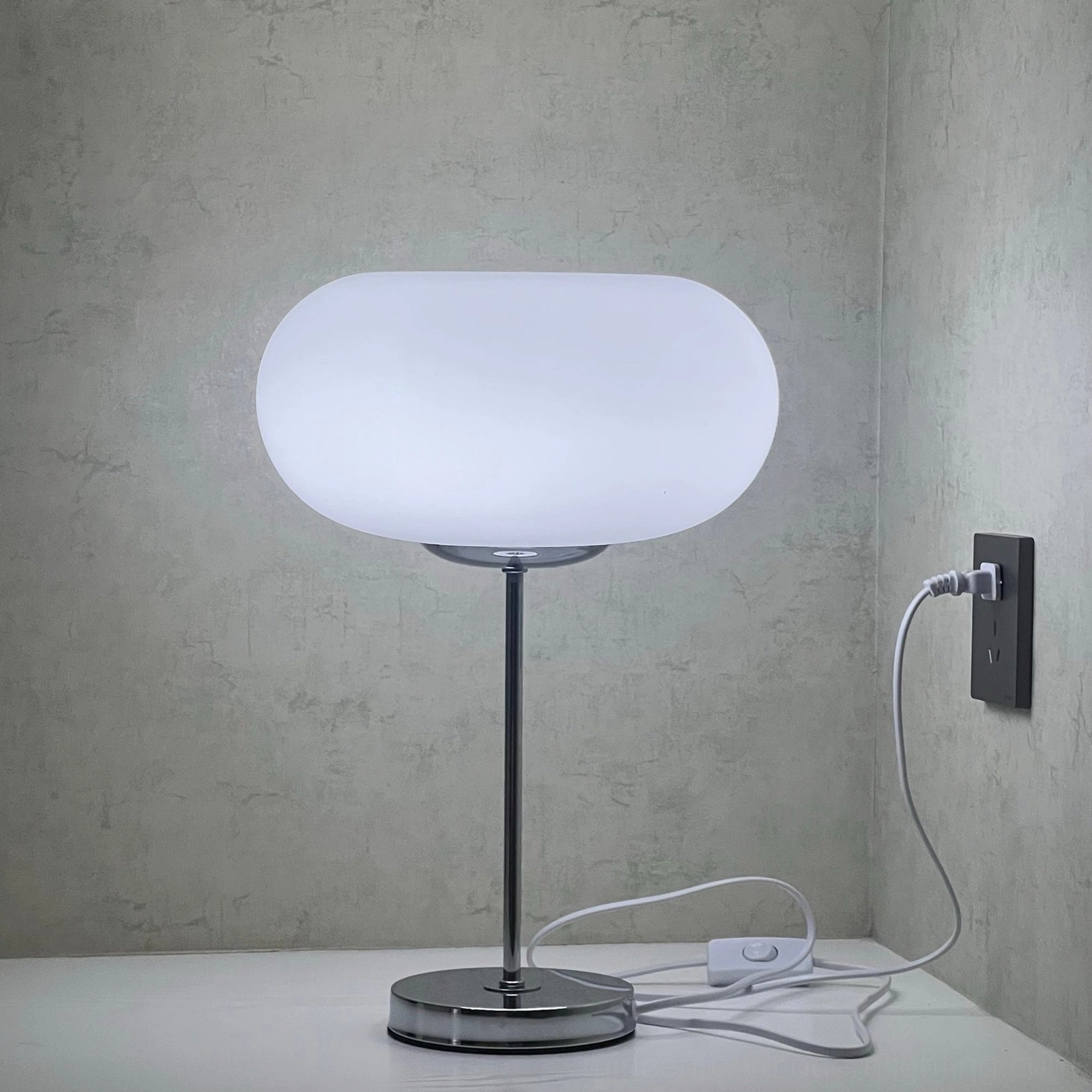 Modern minimalist table lamp with silver metal base and on-off push button switch detail, concrete wall backdrop