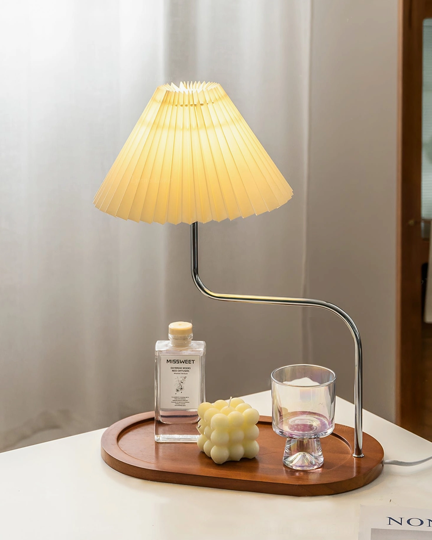 Side view of modern pleated lamp on table near window with skincare bottle and decorative glass cup