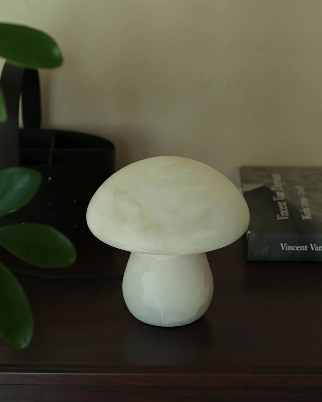 Compact mushroom alabaster table lamp with soft neutral finish beside green houseplants on dark wooden surface