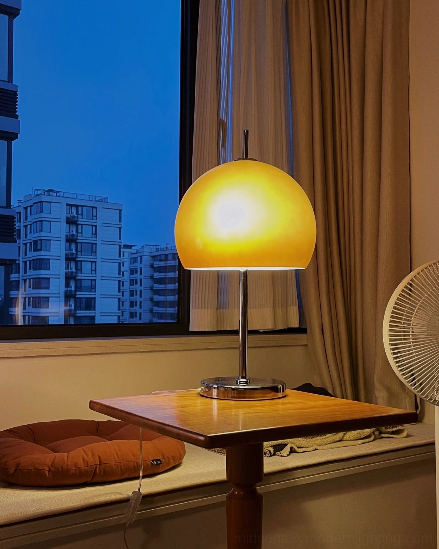 Evening view of lit mushroom table lamp with warm yellow shade on wooden side table near window