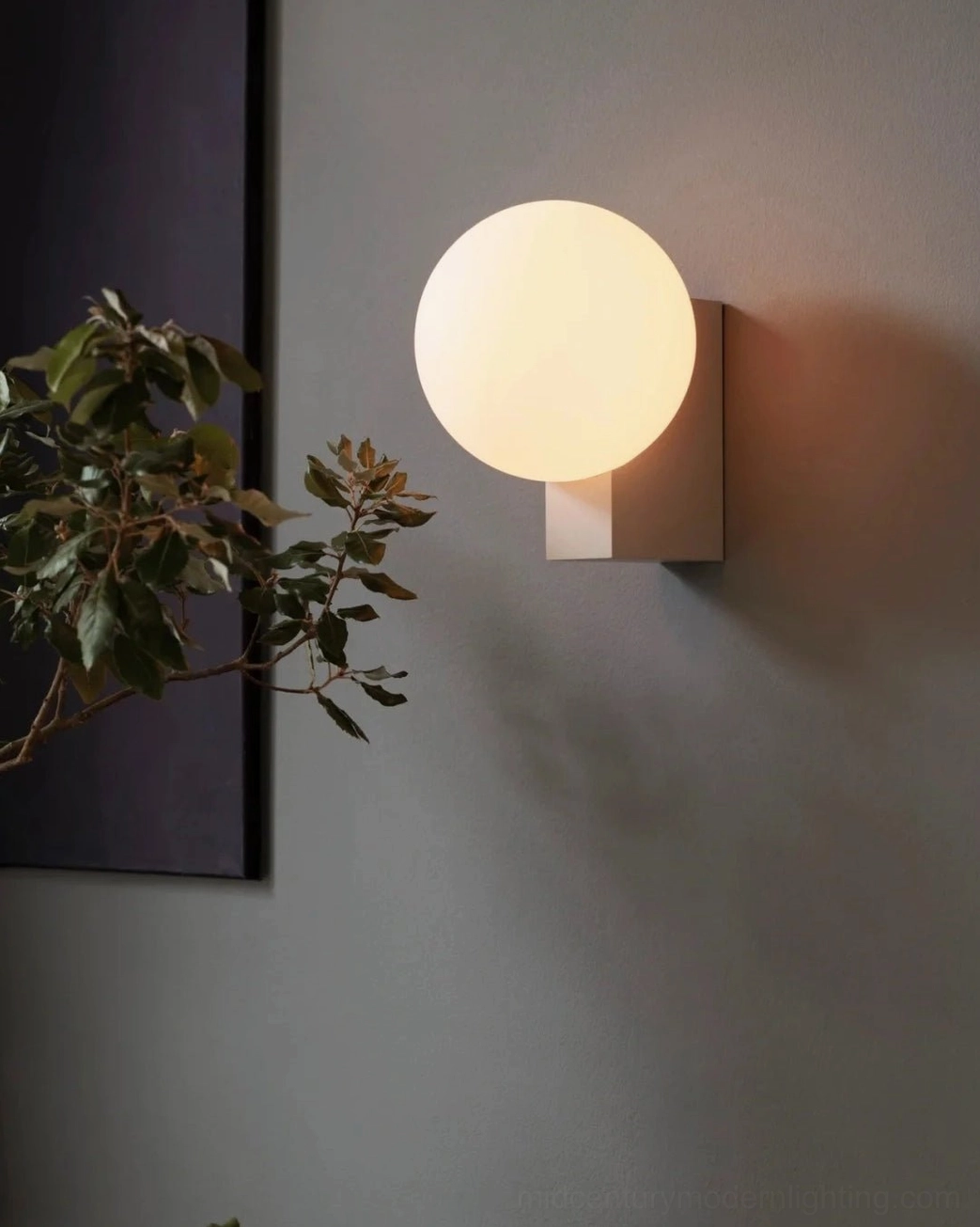 Modern globe wall sconce with off-white metal base creating soft lighting against wall with artwork and foliage