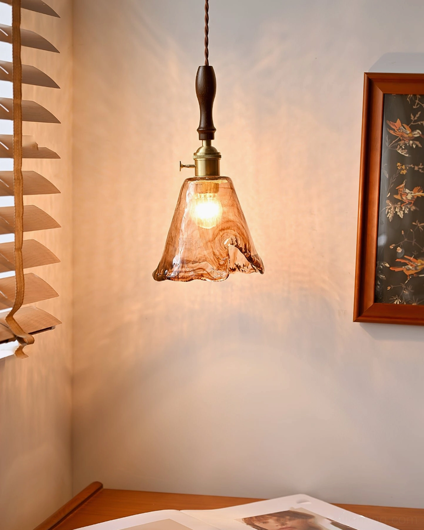 Organic glass pendant lamp with glowing bulb in sunlit corner near window blinds, neutral home decor