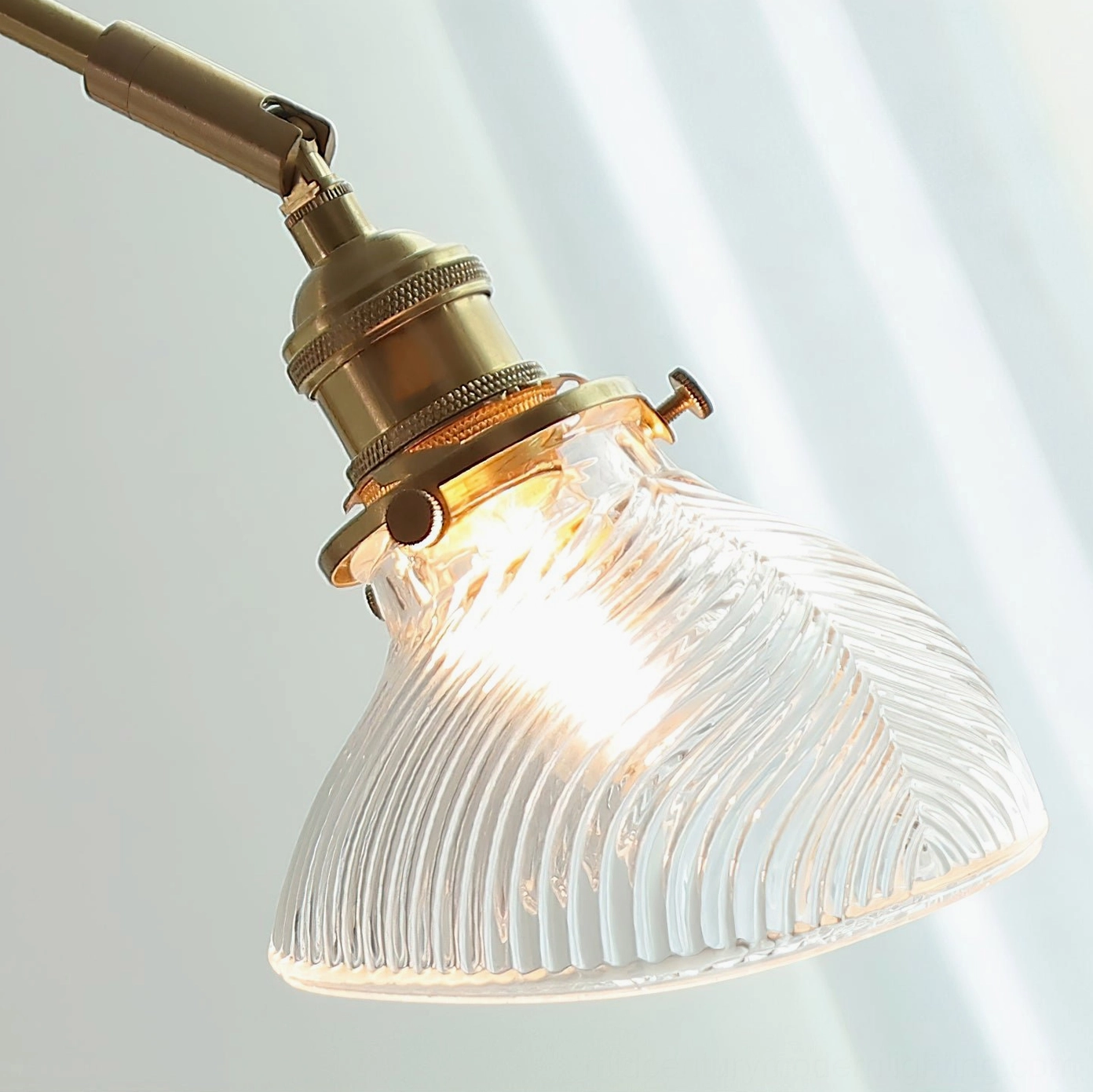 Rebell swing arm lamp close-up side angle showing brass joint, clear ribbed glass shade, and exposed bulb detail on striped background