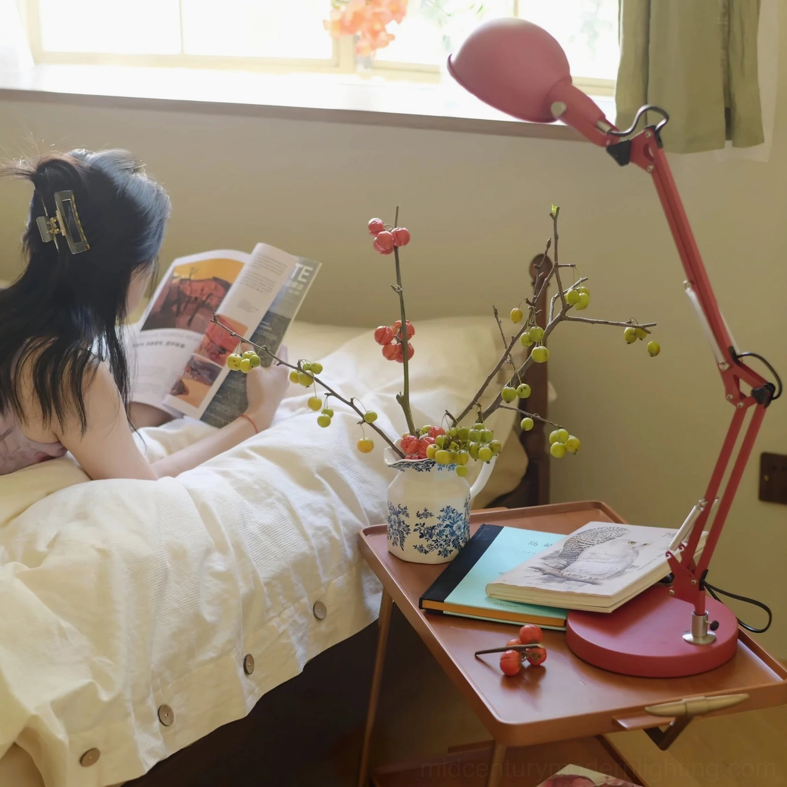Woman reading in bed under red swing arm table lamp with floral decor and books on bedside table