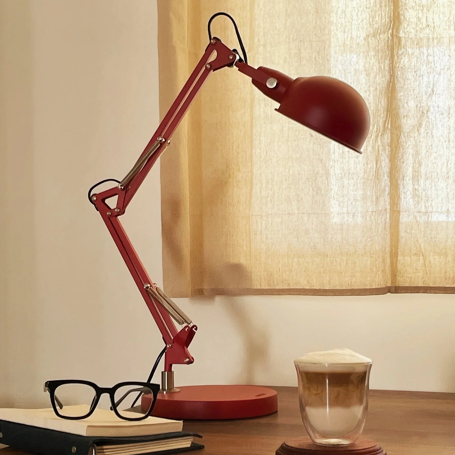 Red swing arm desk lamp on wooden desk close-up with reading glasses, coffee cup, and sheer curtain backdrop