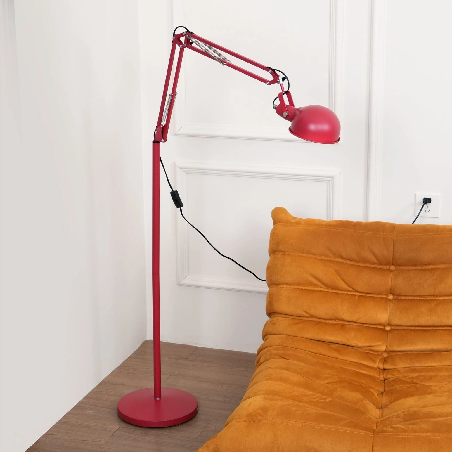 Red adjustable floor lamp in modern living room beside orange sofa, displaying full swing arm extension and black power cord