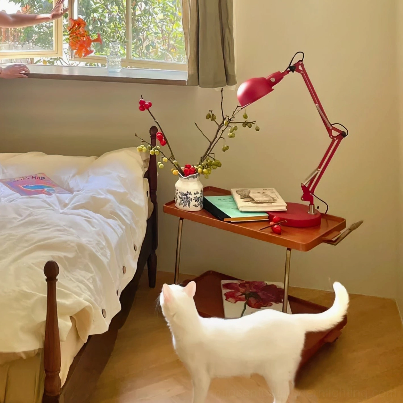 Red swing arm table lamp in bedroom setting with white cat nearby and floral side table arrangement