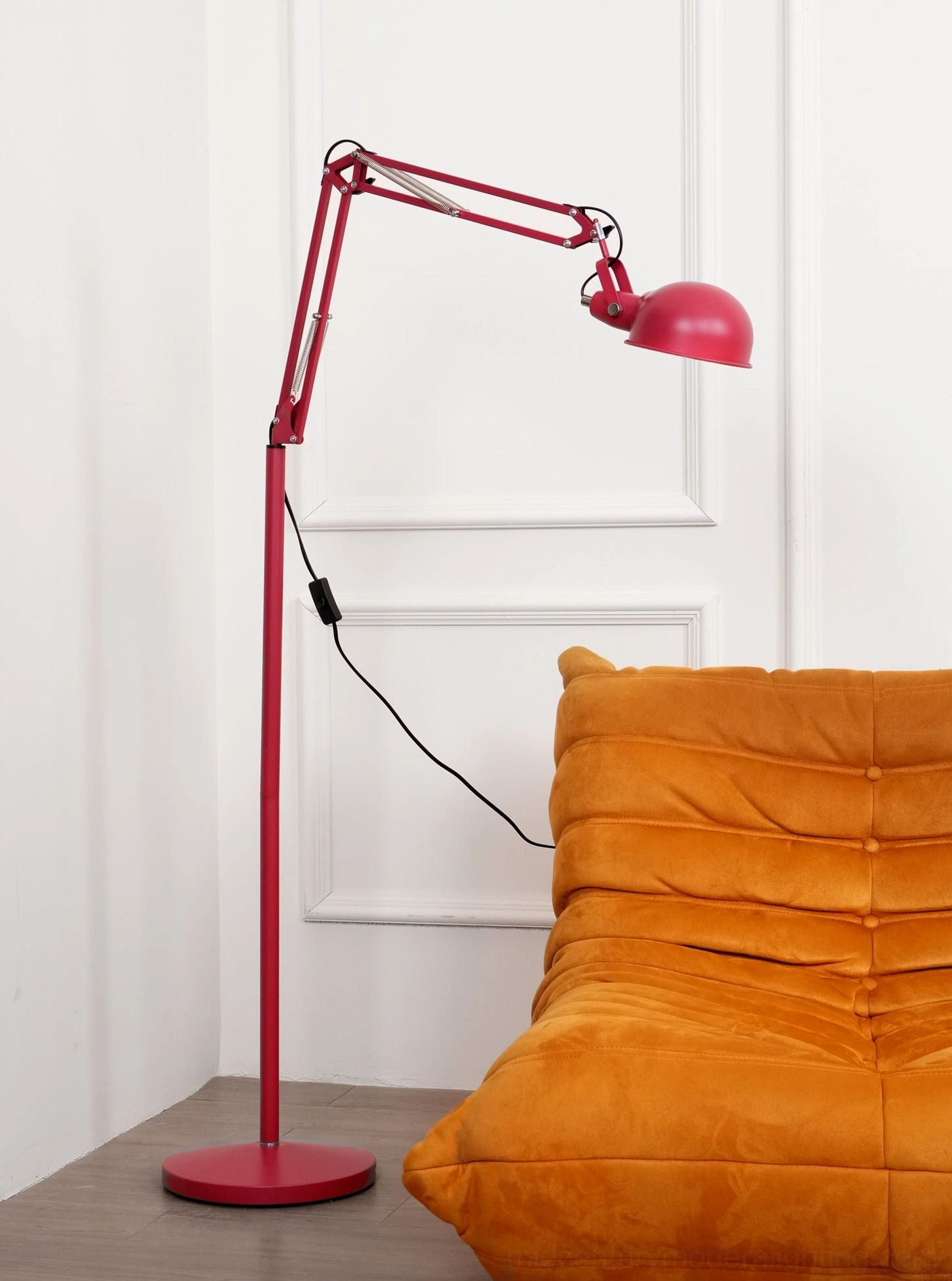 Red adjustable floor lamp positioned in living room interior, side angle showing articulated arm and matte red finish