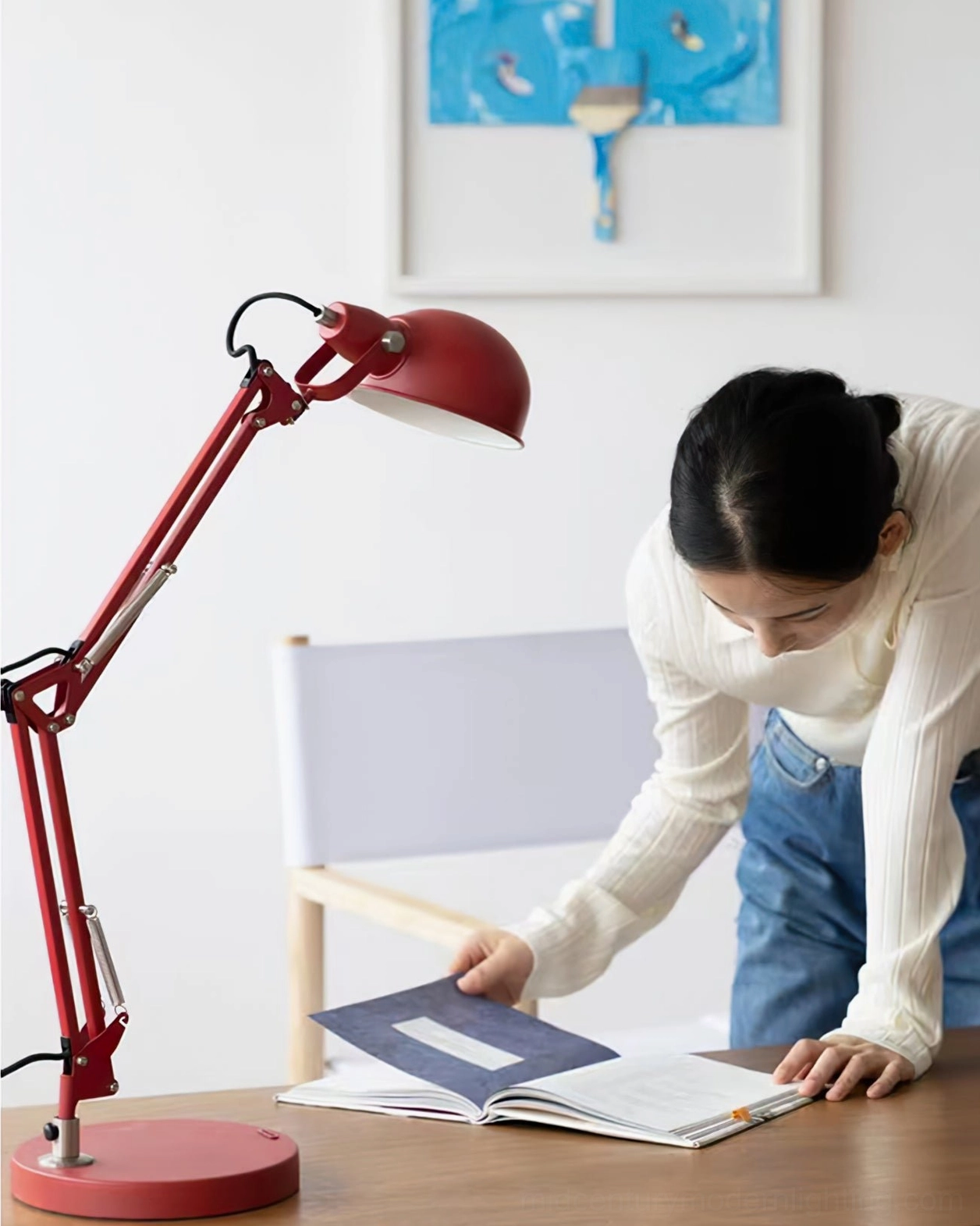 Woman in white top and blue jeans reading under red adjustable desk lamp at home study desk