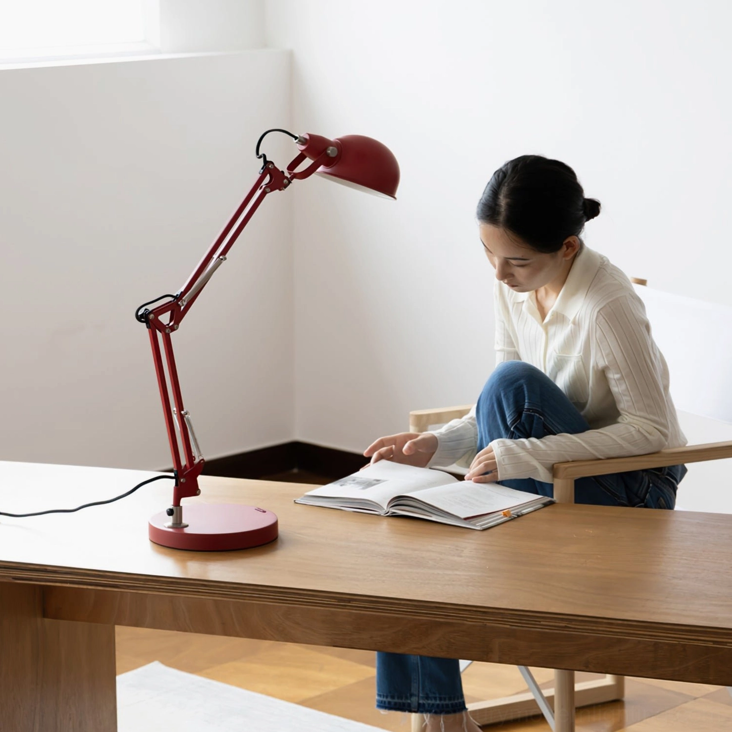 Woman in white knitwear reading book beside red swing arm desk lamp at wooden home office desk