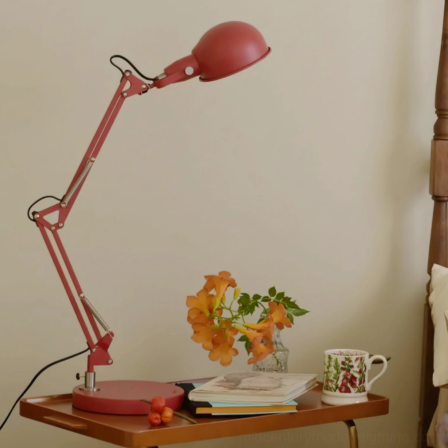 Red adjustable desk lamp on wooden side table with dried flowers, ceramic cup, and warm-toned home furnishings