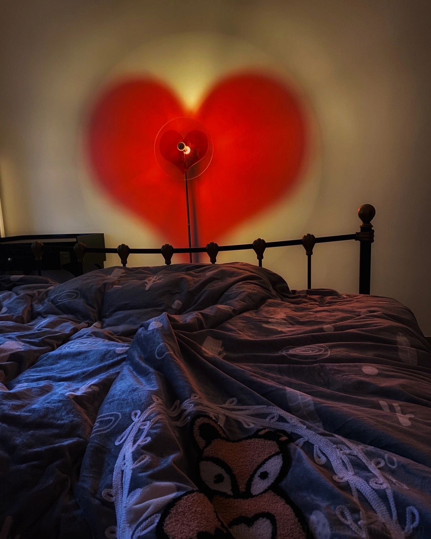 Heart projection floor lamp in cozy bedroom creating soft warm red shadow over patterned bedding, romantic ambiance