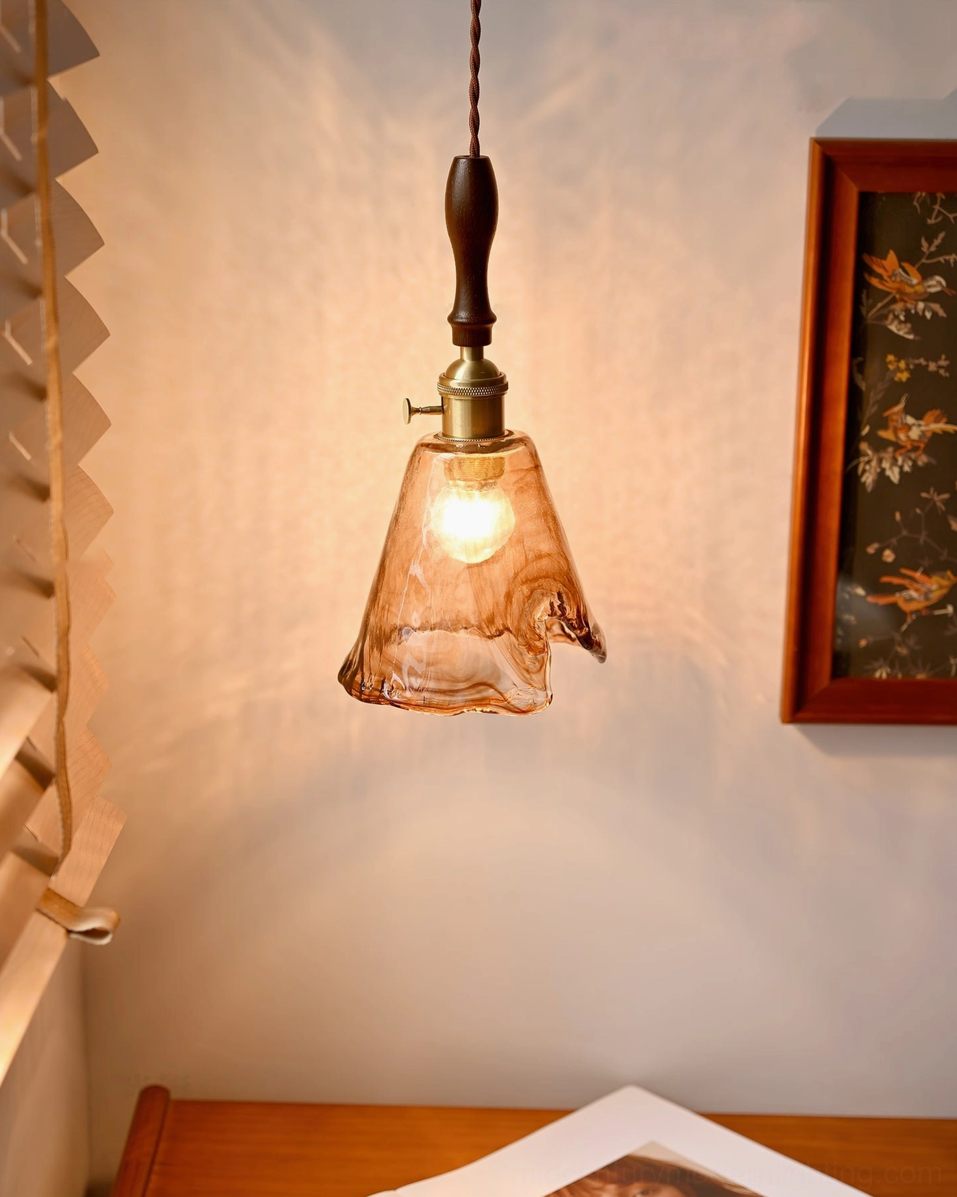 Sculptural pendant lamp with irregular glass form creating warm glow above wooden desk in minimalist room